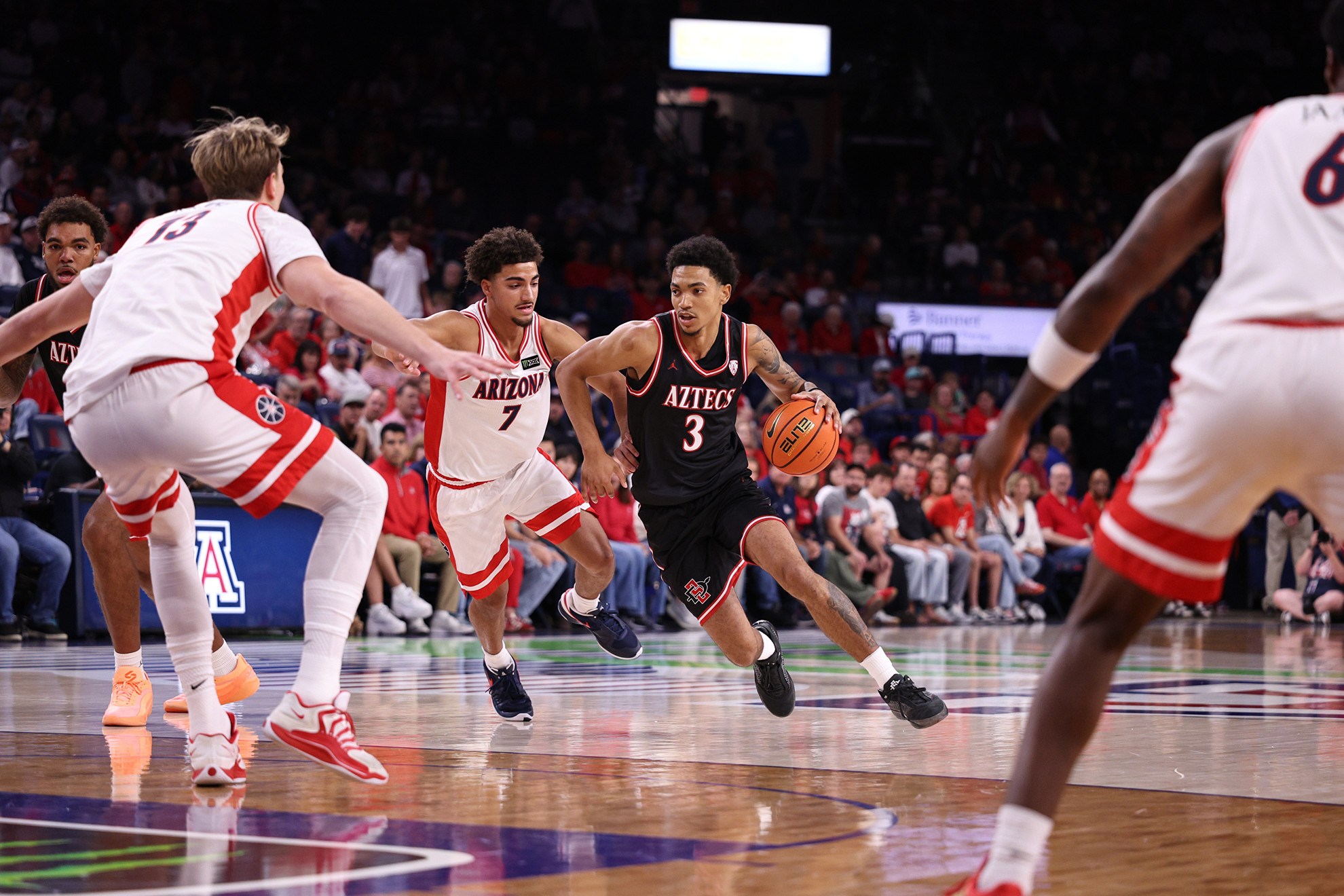 A basketball player in a black jersey dribbles the ball while being guarded by opponents in white and red uniforms during a game.